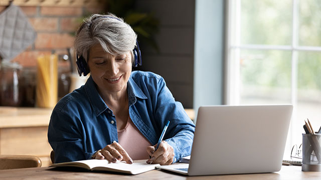 Señora de pelo blanco, con auriculares, tomando nota en un cuaderno de lo que escucha en la computadora a que tiene enfrente